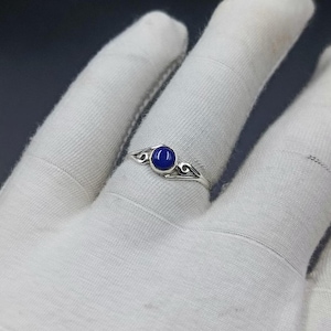 Puede incluir: Un anillo de plata con una pequeña piedra cabujón redonda de color azul intenso engastada en el centro. La banda está elaborada con delicados patrones de metal en espiral que se extienden desde el engaste a lo largo de la fina banda.
