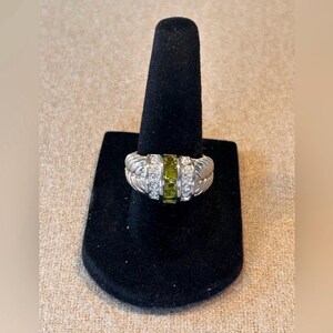 May include: A silver ring with a central row of green stones flanked by rows of clear stones. The ring has a textured design on the sides and is displayed on a black ring holder.