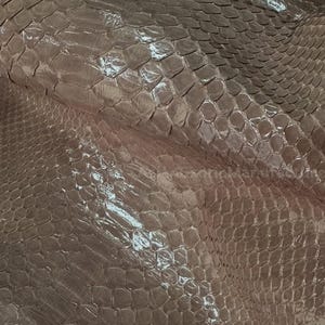 May include: Close-up of a sheet of pinkish-brown snakeskin leather. The scales are visible and have a glossy finish. The leather is folded and appears to be a large piece. The texture is detailed and the color is consistent throughout.