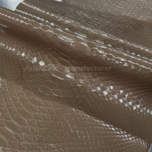 May include: Close-up of a sheet of tan-coloured, textured leather with a reptile-like scale pattern. The leather is folded over, revealing its underside. Rolled leather pieces are visible in the background. The material has a glossy finish.