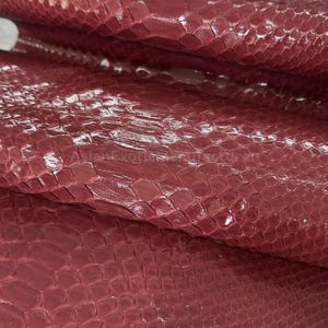 Pode incluir: Close-up de uma folha de couro de pele de cobra bordô. O couro tem um acabamento brilhante e um padrão de escamas. O material está dobrado, revelando o verso e a textura. Este couro é provavelmente usado para artesanato.
