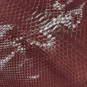 May include: Close-up of a burgundy snakeskin leather. The leather has a glossy finish and a scale pattern. The material is rolled up, showing the texture and colour variations. Suitable for various crafting projects.