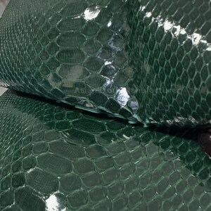 Pode incluir: Close-up de couro de pele de cobra verde escuro com um padrão de escamas. O couro tem um acabamento brilhante e é dobrado, revelando a parte inferior. A textura é detalhada e a cor é consistente em todo o material.