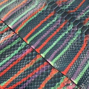 Könnte beinhalten: Nahaufnahme von mehreren Lederstücken mit Schlangenmuster. Das Leder ist in einem dunklen Grauton mit diagonalen Streifen in Orange, Grün und Lila gefärbt. Die Textur des Leders ist sichtbar.