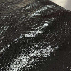Pode incluir: Close-up de um material preto texturizado com um padrão semelhante ao de um réptil. A superfície reflete a luz, destacando as escamas e criando uma aparência brilhante. O material parece ser couro ou um tecido semelhante.
