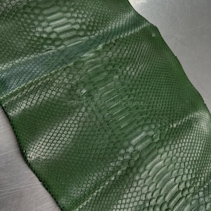 Op de afbeelding: Close-up van een stuk groen slangenleer. Het leer heeft een getextureerd, geschubd patroon met verschillende groentinten. De schubben zijn klein en dicht opeengepakt, waardoor een gedetailleerd oppervlak ontstaat.