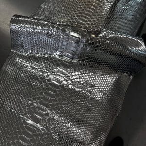 Op de afbeelding: Close-up van een zilveren slangenleer materiaal. Het materiaal heeft een textuur, schubachtig patroon met een metallic glans. Het leer is omgevouwen, waardoor de onderkant zichtbaar wordt en de textuur wordt benadrukt.