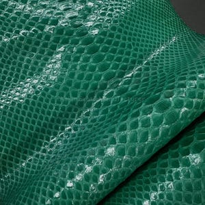 Pode incluir: Close-up de uma folha de couro de pele de cobra verde esmeralda. O couro tem um padrão texturizado, semelhante a escamas, com um acabamento brilhante. As dobras do couro são visíveis, criando sombras e destaques que enfatizam a textura.