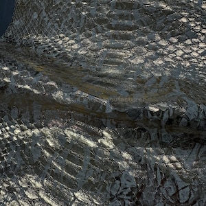 Pode incluir: Close-up de um material texturizado com aparência metálica e padrão escamoso. A superfície tem um brilho escuro e iridescente com destaques e sombras, sugerindo pele de réptil ou textura semelhante. A cor geral é uma mistura de cinza escuro e prata.