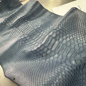 Pode incluir: Close-up de um pedaço de couro de pele de cobra azul-acinzentado. O couro apresenta um padrão texturizado e escamoso com variações de cor e tom. As escamas são visíveis e criam um design natural único. O couro está dobrado, mostrando sua flexibilidade.