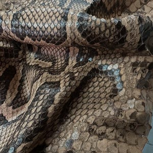 May include: Detailed image of snakeskin texture. The leather shows a pattern of tan, brown, and black scales, giving a natural, reptilian look. The scales vary in size and shape, adding depth and detail to the material.