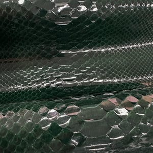 Pode incluir: Close-up de pele de cobra verde escuro com acabamento brilhante. As escamas são visíveis e criam um padrão texturizado. A imagem mostra as variações naturais de textura e cor do material.