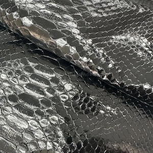 Peut inclure: Gros plan sur une feuille de similicuir noir brillant avec un motif peau de serpent. Le matériau a une surface texturée avec un motif d'écailles répétitif, reflétant la lumière et créant des reflets et des ombres. L'effet global est une texture sombre et luxueuse.