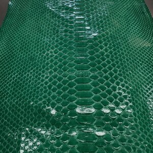 May include: Close-up of a sheet of emerald green snakeskin leather. The leather has a textured, scaly pattern with a glossy finish. The scales are arranged in a repeating, overlapping design.