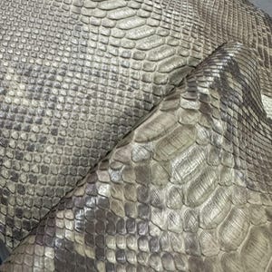 Pode incluir: Close-up de um padrão de pele de cobra texturizado em tons de cinza e marrom. As escamas variam em tamanho e forma, criando uma textura natural e orgânica. A paleta de cores inclui tons de cinza, marrom e toques de branco, dando-lhe uma aparência realista. Adequado para vários projetos de artesanato.