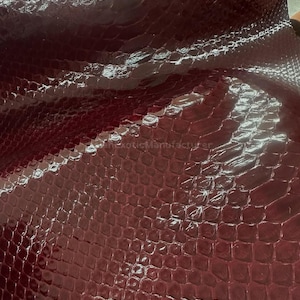 Pode incluir: Close-up de uma folha de couro de pele de cobra na cor bordô. O couro tem um acabamento brilhante e um padrão de escamas. A textura é detalhada, com cada escama claramente definida. A aparência geral é rica e luxuosa, adequada para vários projetos de artesanato.