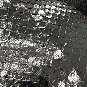 Puede incluir: Primer plano de un material texturizado de color gris oscuro con un patrón similar a escamas. Las escamas tienen un brillo metálico reflectante, creando reflejos y sombras. El efecto general es una superficie texturizada e iridiscente.