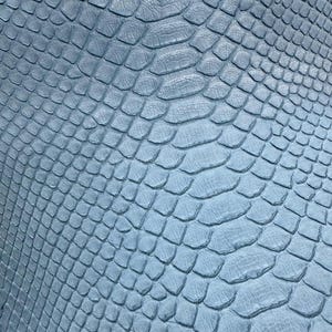 May include: A close-up view of a light blue snakeskin pattern. The textured surface shows a repeating scale design, with each scale slightly raised and overlapping. The color is a soft, muted blue, creating a subtle, elegant appearance. This material could be used for fashion or craft projects.