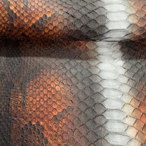 Pode incluir: Close-up de um padrão de pele de cobra. As escamas são uma mistura de castanho, laranja, cinza e branco, criando uma superfície texturizada. O padrão é detalhado e parece natural, com variações de cor e tamanho.