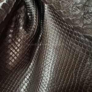 May include: Close-up of a dark gray snakeskin pattern. The material has a textured, scaly appearance with a subtle sheen. The scales are arranged in a repeating diamond pattern, creating a luxurious and exotic look. The fabric is folded, showing depth and dimension.