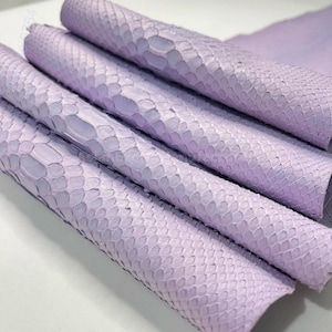 Pode incluir: Rolos de couro de pele de cobra de cor lavanda. O couro apresenta um padrão de escamas e está disposto na diagonal. O material parece macio e flexível, adequado para vários projetos de artesanato.