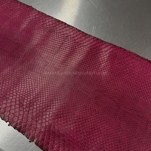 May include: A close-up of a piece of burgundy-colored snakeskin leather. The leather has a textured, scale-like pattern. The material is likely used for crafting items like wallets, belts, or other accessories.