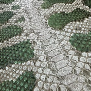 Peut inclure: Gros plan sur un motif peau de serpent dans les tons de vert, gris et blanc. La texture est détaillée, avec des écailles et un motif camouflage. Le motif est probablement utilisé pour des accessoires de mode ou de la sellerie.