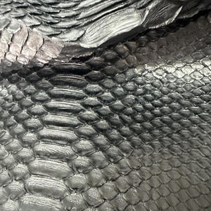 Puede incluir: Primer plano de un material texturizado gris metálico con un patrón de escamas. La superficie tiene una textura ondulada en algunas zonas, creando un juego de luces y sombras. El efecto general es una apariencia lujosa, similar a la de un reptil.