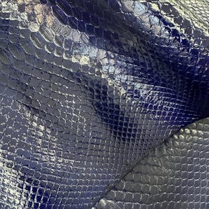 Pode incluir: Close-up de um material azul marinho texturizado com um padrão escamoso. A superfície reflete a luz, criando destaques e sombras. O material parece flexível e possivelmente couro ou um tecido semelhante.
