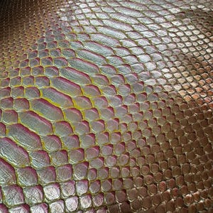 Pode incluir: Close-up de um material texturizado e iridescente com um padrão escamoso. A superfície transita de um tom bronze para prata e rosa, com destaques amarelos. O design assemelha-se à pele de réptil, adequado para projetos de moda ou artesanato.