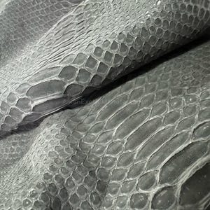 May include: Close-up of a sheet of grey snakeskin leather. The leather has a textured, scaly pattern with varying shades of grey. The scales are tightly packed and create a unique, natural design. The image highlights the texture and detail of the exotic material.