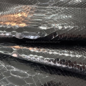 May include: Close-up of black snakeskin leather. The leather has a glossy finish and a scale pattern. The texture is detailed, with visible ridges and variations in the scales. The image shows the natural texture and sheen of the exotic material.