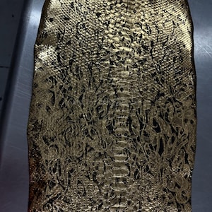Pode incluir: Close-up de um tecido dourado com um padrão texturizado semelhante ao de um réptil. O tecido tem um design escuro sobre a base dourada. O padrão é intrincado e detalhado, assemelhando-se a escamas e formas giratórias.