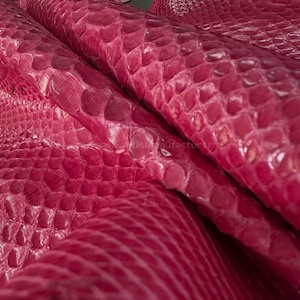 Pode incluir: Close-up de couro de pele de cobra rosa vibrante. O couro tem um padrão escamoso texturizado, com variações de cor e profundidade. A imagem mostra várias camadas do material, destacando sua flexibilidade e textura. A impressão geral é de um material exótico de alta qualidade.