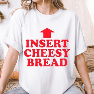 May include: White t-shirt with the text "INSERT CHEESY BREAD" in red, with a red arrow pointing upwards. The shirt is worn by a person sitting down.