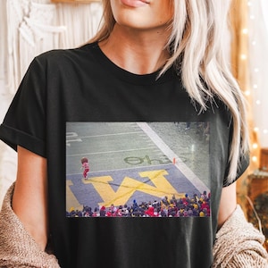 Può includere: T-shirt nero con un'immagine rettangolare di un campo da football, che mostra una grande "M" gialla e una folla. Una persona in abito rosso è sul campo. La maglietta è un capo casual.