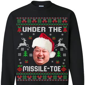May include: Black sweatshirt with a humorous Christmas design. The design features a face wearing a Santa hat, with the text "UNDER THE MISSILE-TOE" in white. The design also includes reindeer, bells, and snowflakes in red and green.