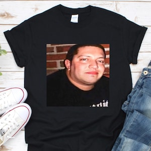 May include: Black t-shirt with a printed image of a person's face. The image is in a square shape and is centered on the shirt. The shirt is paired with blue jeans and white sneakers.