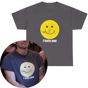 Peut inclure: T-shirt gris foncé avec un smiley jaune vieilli et le texte "I hate you" en dessous. Le graphisme a un aspect vintage et usé. Le t-shirt est de style col rond.