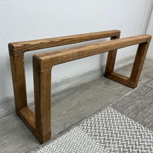 Puede incluir: Un par de taburetes de madera con acabado natural. Los taburetes tienen un marco rectangular con patas anguladas y están colocados sobre un suelo gris junto a una alfombra estampada. La veta de la madera es visible.