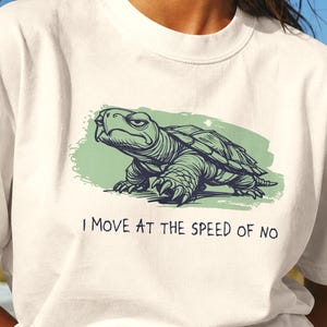 May include: An ivory short-sleeve t-shirt featuring a graphic illustration of a green turtle with a grumpy expression. Below the turtle, the text reads, "I MOVE AT THE SPEED OF NO" in black capital letters. The design is set against a light green brushstroke.