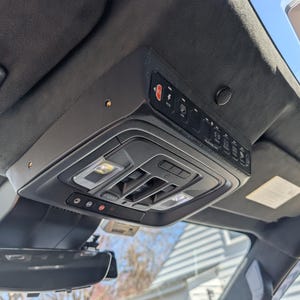 May include: Interior car roof console with various buttons and controls. The console is black and has a built-in light, buttons for lights, and other functions. A rearview mirror and sun visor are also visible.