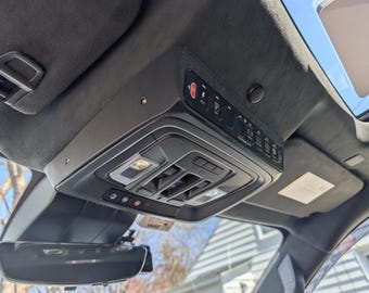 May include: A black overhead console mounted to a dark gray suede-like vehicle headliner. The console features an integrated Auxbeam switch panel with multiple illuminated icons, interior dome lights, and various control buttons for vehicle functions.