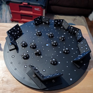 May include: A black game board with white polka dots and several black dice with white dots. The board has several black, L-shaped pieces with white dots around the perimeter. The game is on a wooden table.