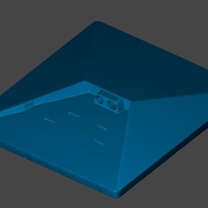 Puede incluir: Una base angular de color turquesa con un área empotrada y varios agujeros pequeños. La base tiene una forma cuadrada plana con bordes biselados y se asienta sobre una superficie gris. El diseño es simple y geométrico.