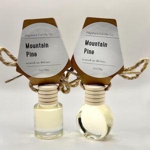 May include: Two glass car diffusers containing light-coloured liquid, each with a wooden cap and a brown cardboard tag. The white labels read Haystack Candle Co, Mountain Pine, scented car diffuser, and 1oz/28g. Each bottle has a tan rope attached.