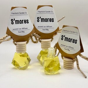May include: Three glass car diffusers with faceted geometric shapes filled with yellow liquid. Each has a wooden cap and a brown paper tag labeled Haystack Candle Co. S'mores scented car diffuser 1oz/28g, attached with gold string.