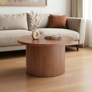 May include: A round wooden coffee table with a brown finish. The table has a cylindrical base with vertical grooves. A vase with white flowers, a book, and glasses are on the table. The table is in front of a beige sofa.