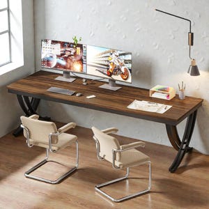 May include: A large, dark wood desk with black metal legs, two computer monitors, and two beige chairs. The desk has a keyboard, mouse, and office supplies. A wall-mounted lamp and a window are visible in the background.