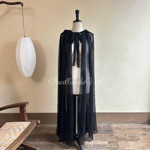 May include: A long, black sheer cape with a hood and tie closure, displayed on a mannequin. The cape is floor-length and made of a lightweight, flowing fabric. The text "Pavillon de Veil" is visible on the cape.
